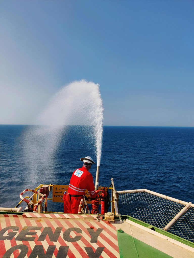 A person in safety gear uses a fire hose on an offshore platform against a sea backdrop.